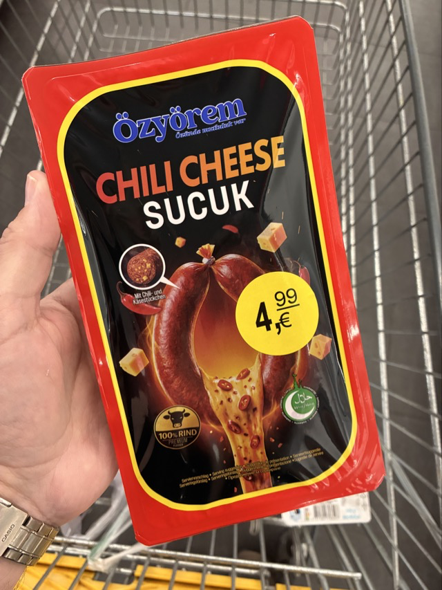 Eine Hand hält eine Packung "Özyörem Chili Cheese Sucuk". Im Hintergrund ein Einkaufswagen.